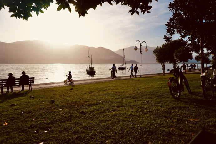 Lido erboso al tramonto con persone sedute su panchina e bimbi in bicicletta. Lago e montagne in secondo piano